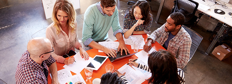 Differing cultural norms, values, and beliefs between two merging companies will inevitably come into conflict. Photo of a team collaborating around an orange desk
